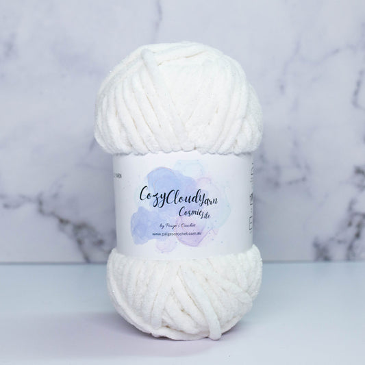 A skein of white Cozy Cloud Cosmic Lite yarn with a label, displayed against a marble background. It is a micro polyester yarn with a slight sparkle to it, approx 5mm in thickness