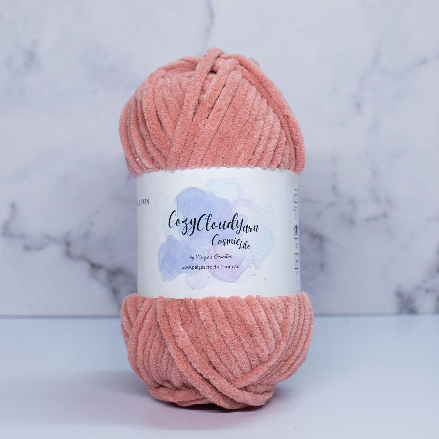 A skein of Cozy Cloud Cosmic Lite yarn with a label, displayed against a marble background. It is a micro polyester plush yarn with a slight sparkle to it, approx 5mm in thickness