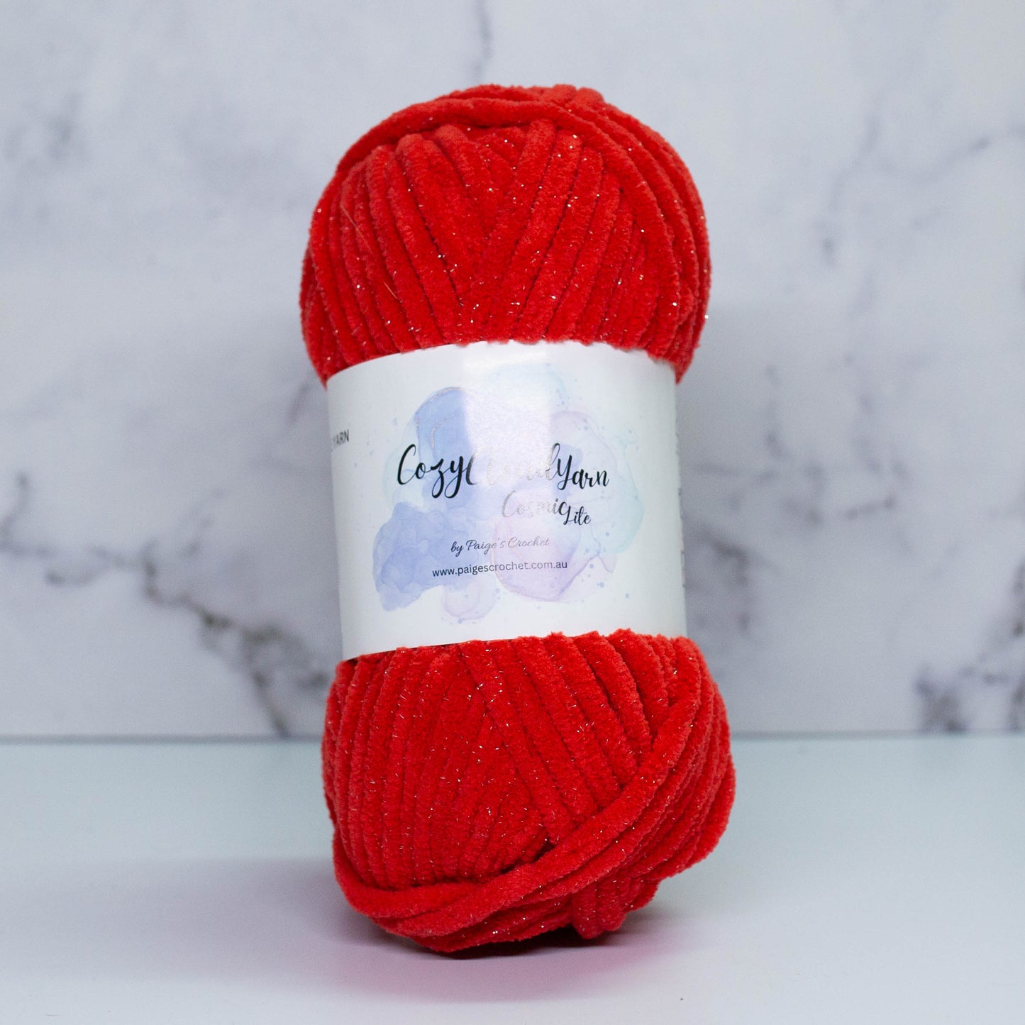 A skein of red Cozy Cloud Cosmic Lite yarn with a label, displayed against a marble background. It is a micro polyester yarn with a slight sparkle to it, approx 5mm in thickness