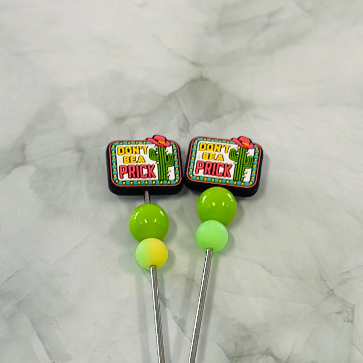 Two novelty stitch sticks with green and yellow ends and colourful 'Don't Be a Prick' text on a marble surface. Amigurumi plushies, sewinf sticks chenille yarn plush yarn