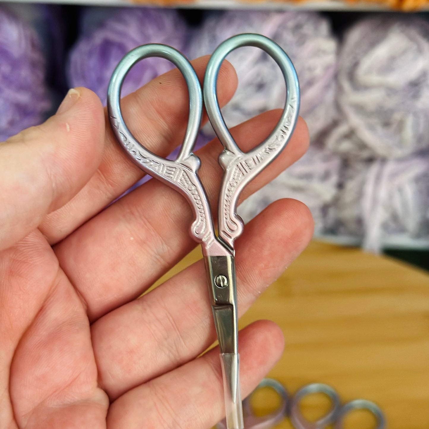 A pair of metal sewing scissors with pastel pink and blue hues, held in a hand with a blurred background of yarn.