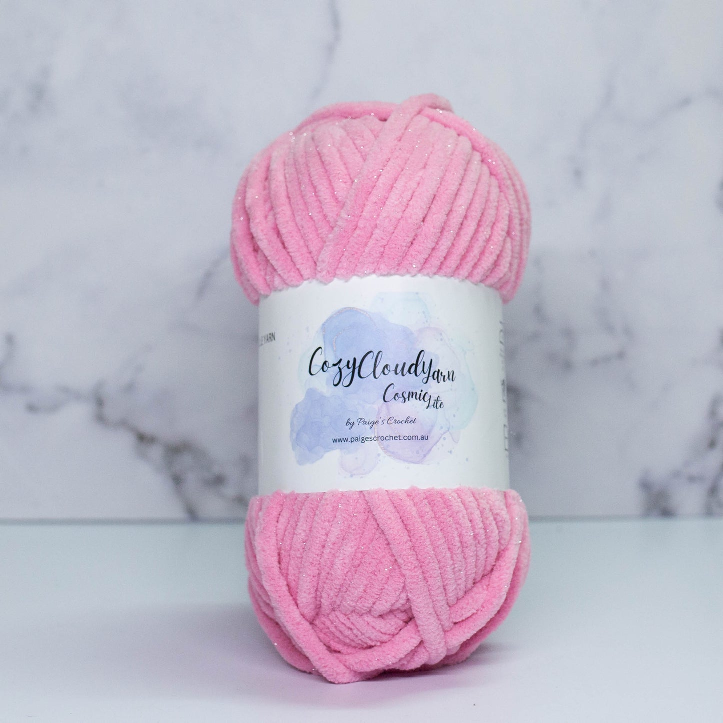 A skein of pink Cozy Cloud Cosmic Lite yarn with a label, placed against a marble background. It is a micro polyester plush yarn with a slight sparkle to it, approx 5mm in thickness