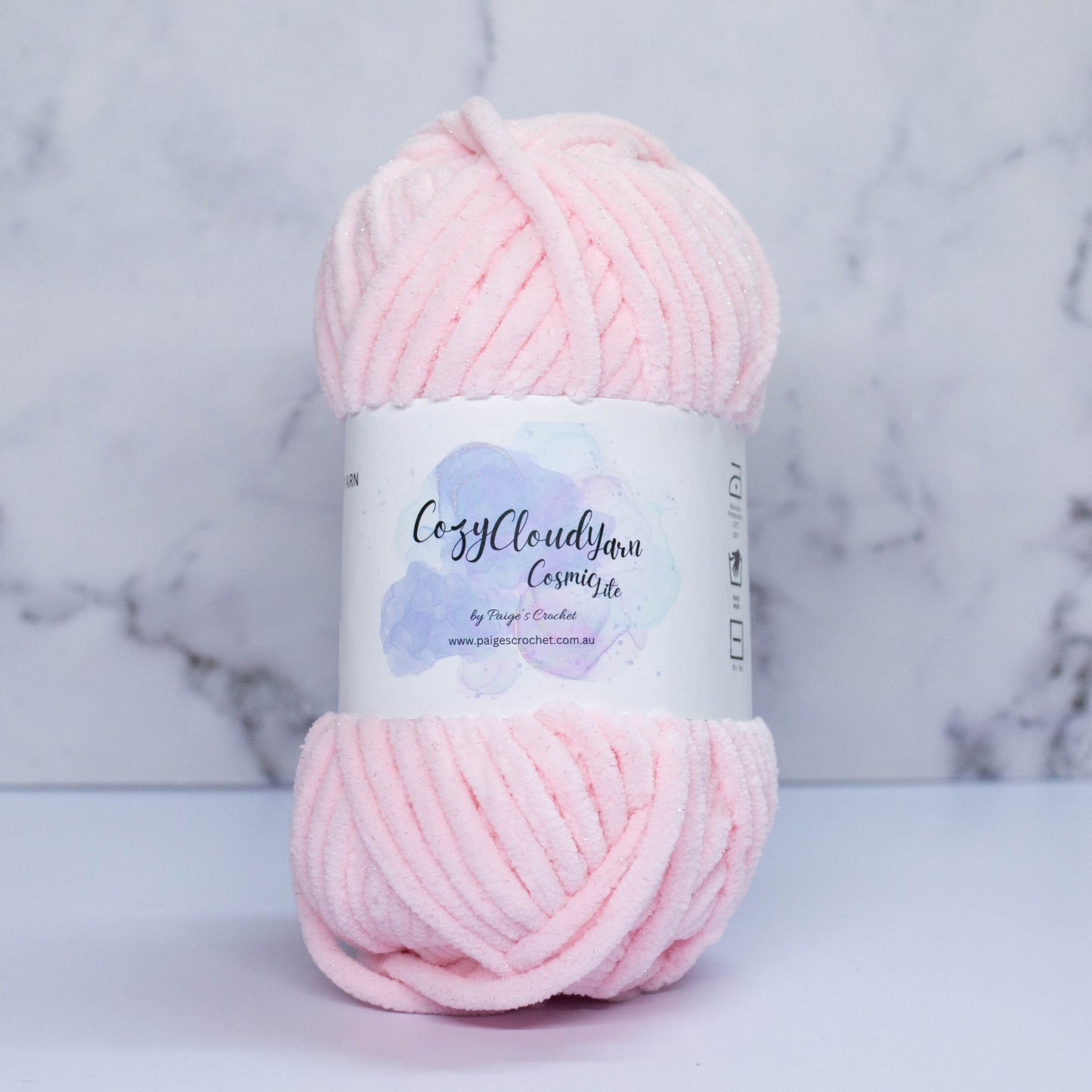 A skein of light pink Cozy Cloud Cosmic Lite yarn with a label, placed against a marble background.It is a micro polyester yarn with a slight sparkle to it, approx 5mm in thickness