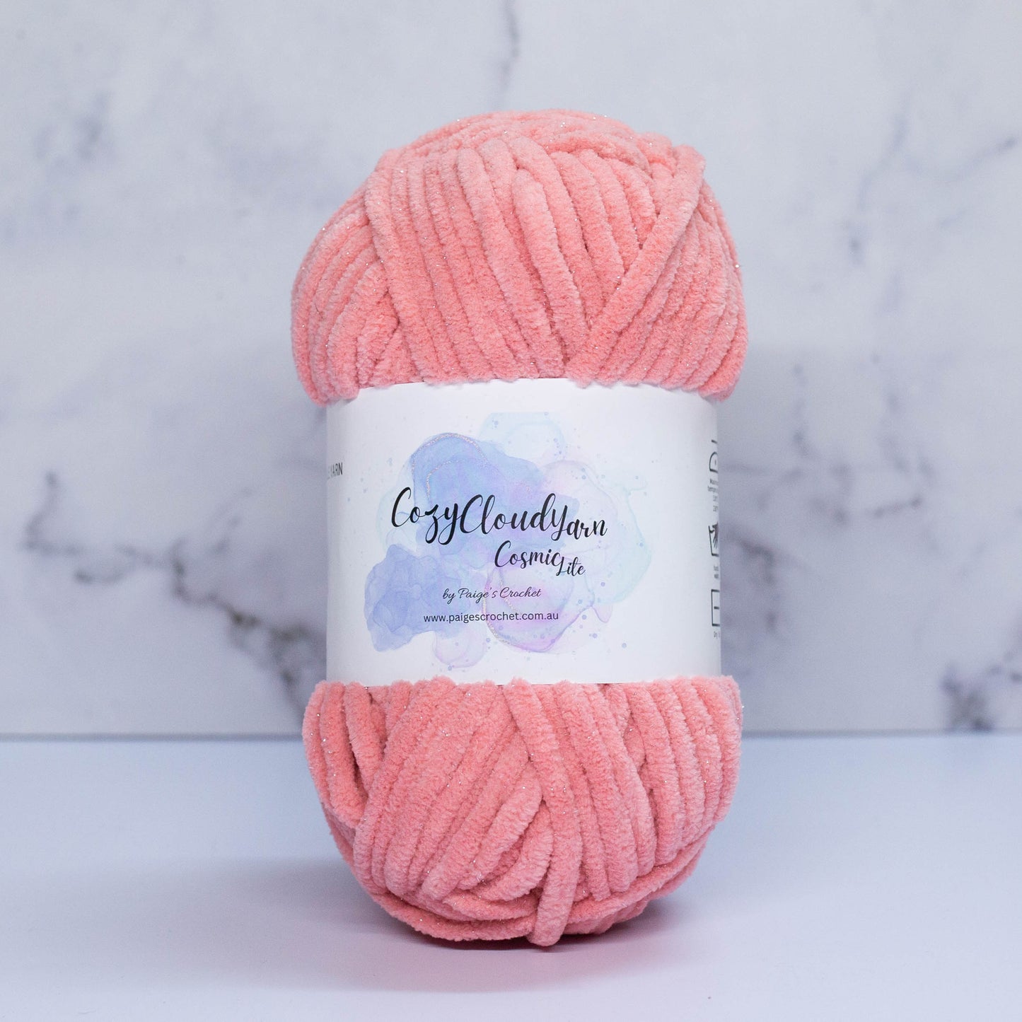 A skein of peachy pink Cozy Cloud Cosmic Lite yarn with a label, displayed against a marble background. It is a micro polyester plush yarn with a slight sparkle to it, approx 5mm in thickness 