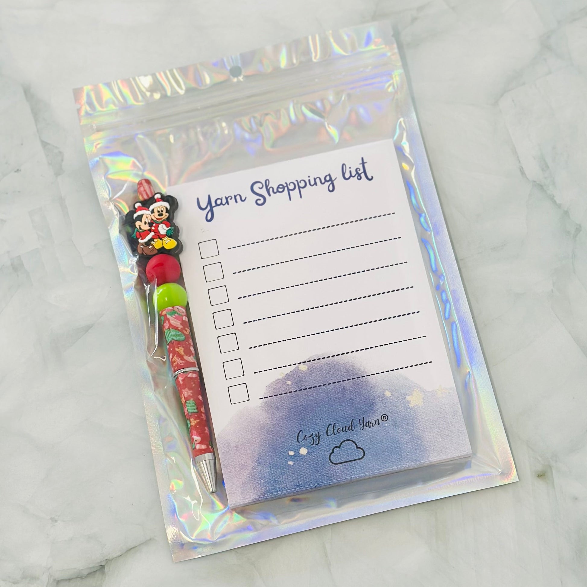 Notepad with 'Yarn Shopping List' and a pen in a holographic case on a marble surface
