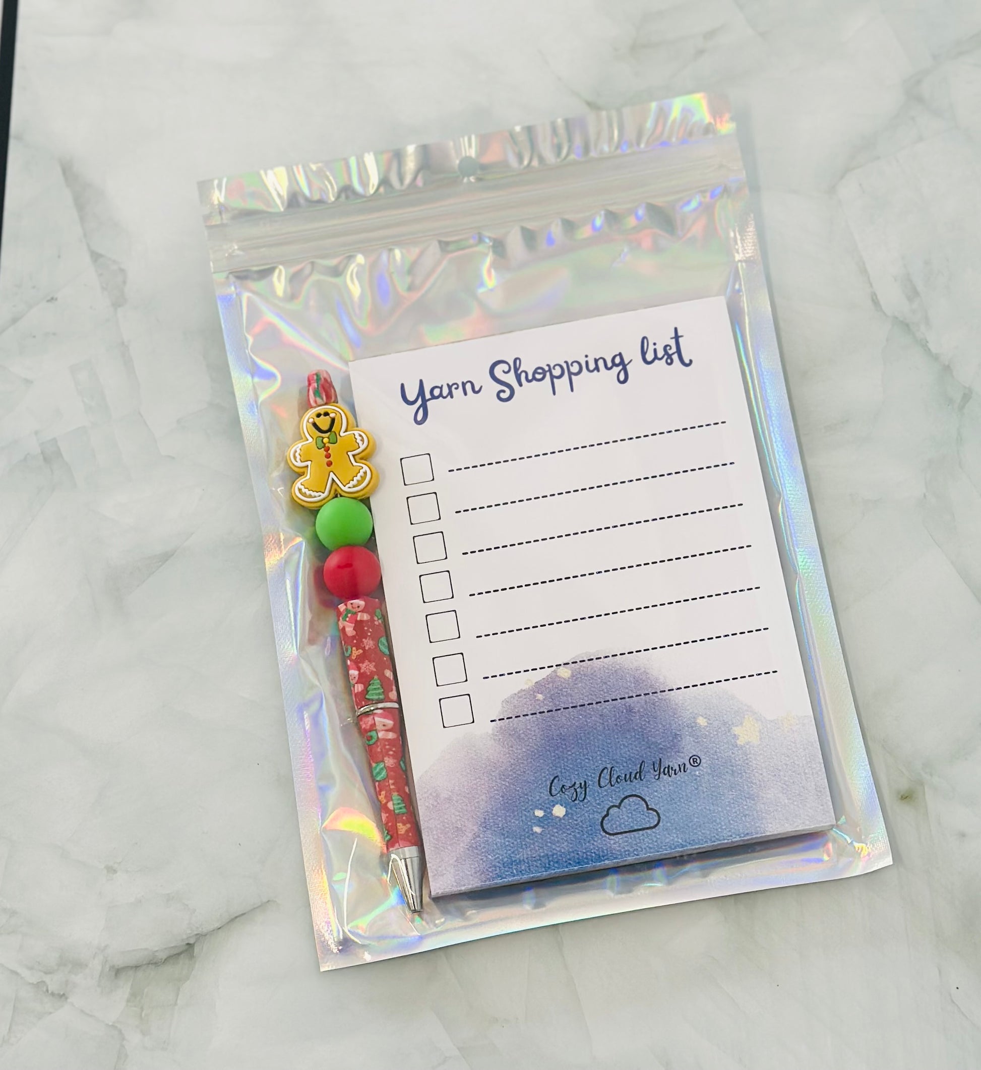 Yarn shopping list with pen and decorative items in a holographic bag on a marble surface