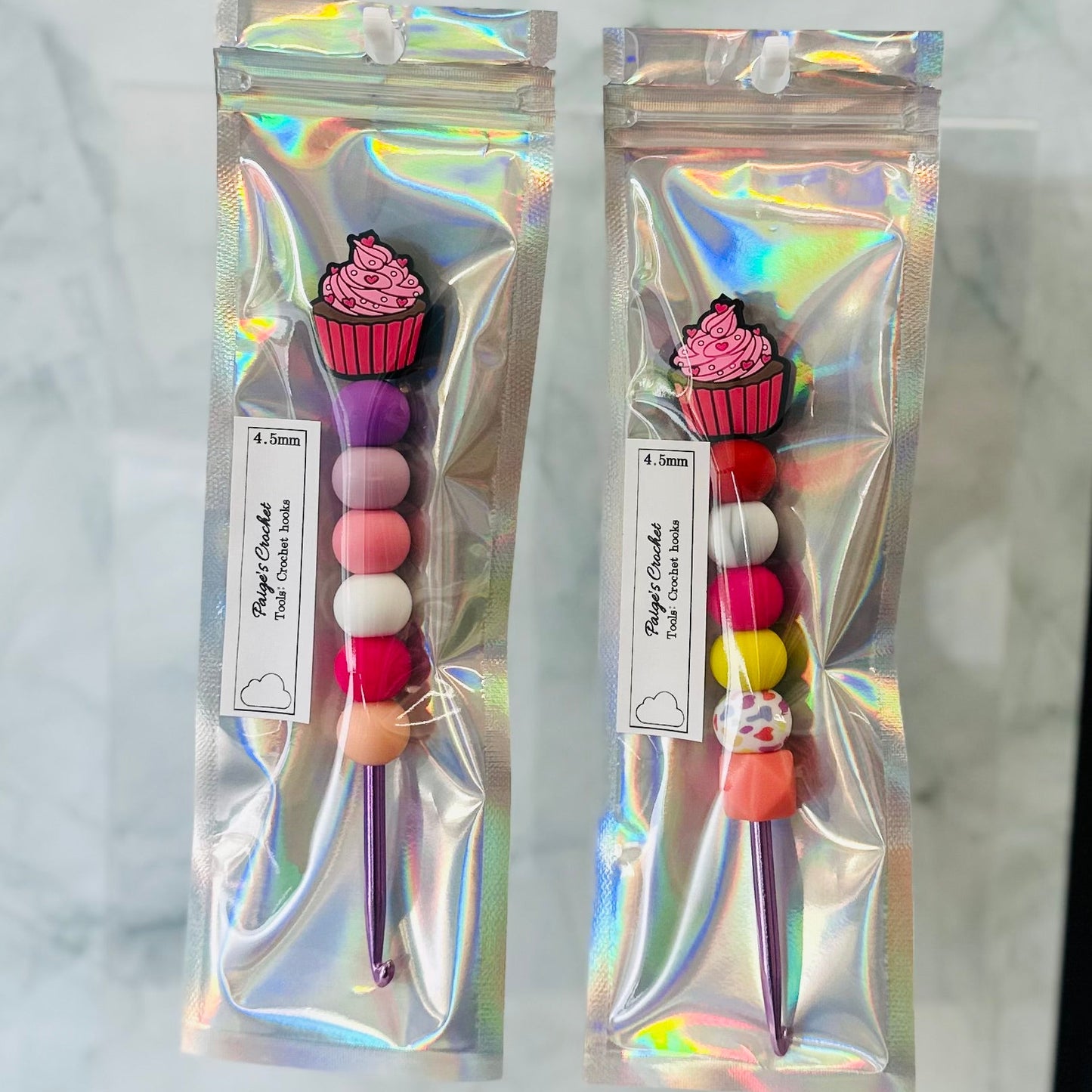 Two colorful crochet hooks accessories in holographic packaging on a marble surface.
